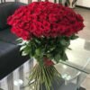 100 Red Roses Grande Bouquet standing on a glass coffee table beside a dark sofa