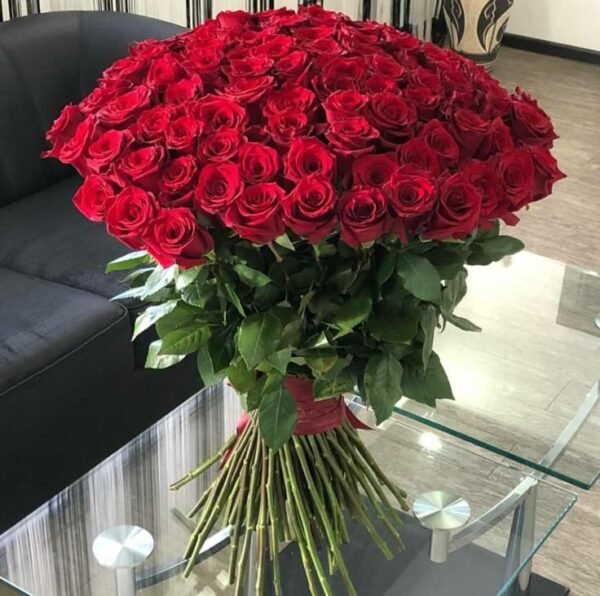 100 Red Roses Grande Bouquet standing on a glass coffee table beside a dark sofa