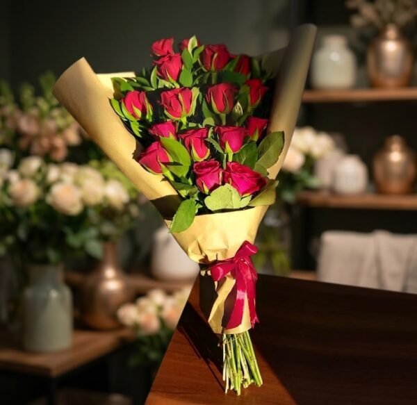 18 Red Roses Bouquet – in a flower shop setting باقة 18 وردة حمراء بورق كرافت موضوعة على طاولة خشبية داخل محل ورد